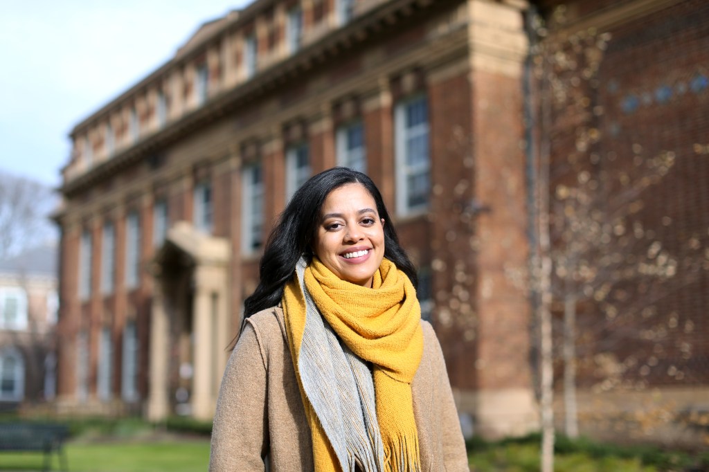 Yalidy Matos wearing a tan coat and large yellow scarf is smiling on Voorhees Mall on College Avenue at Rutgers University.